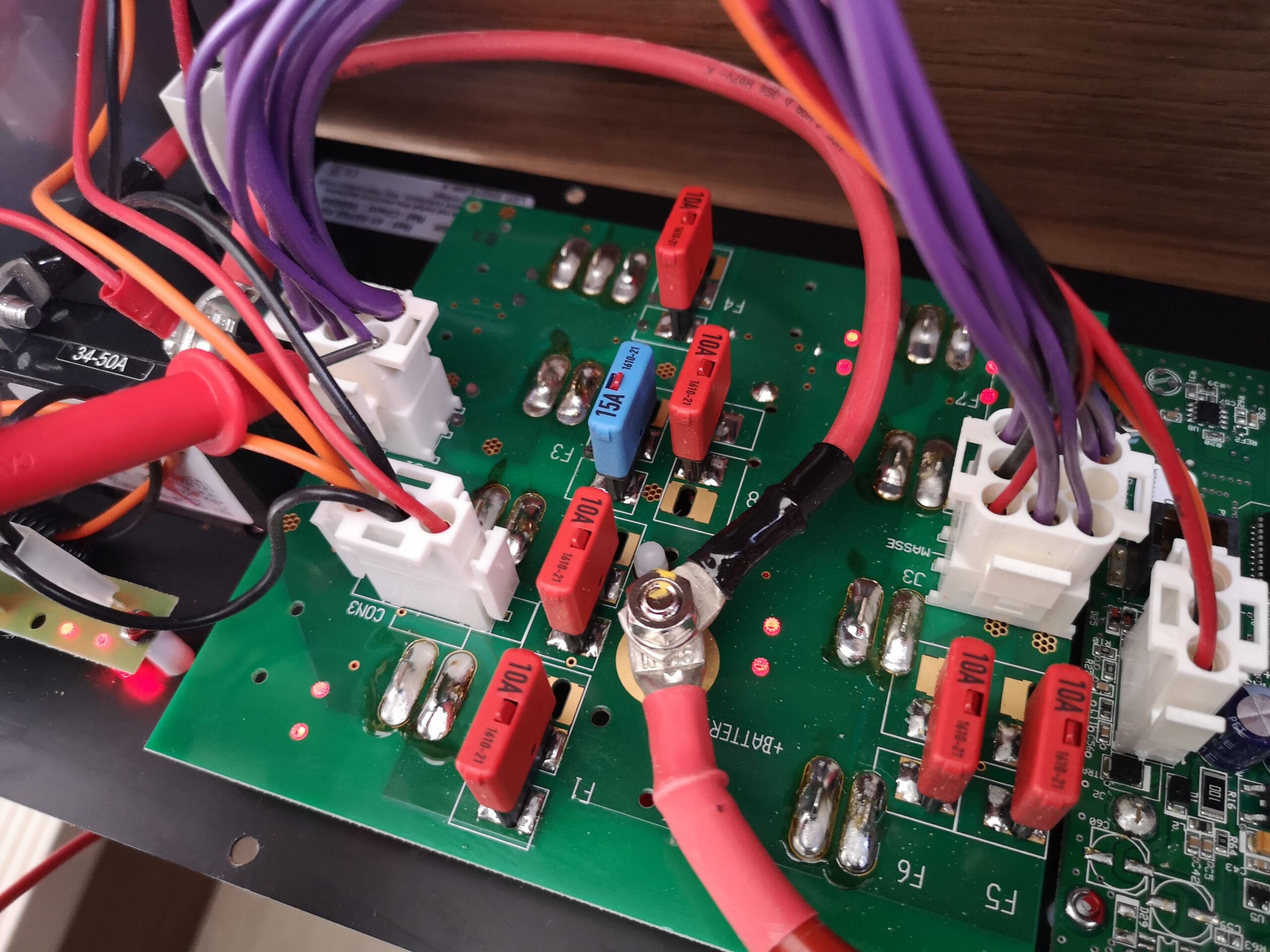 Photo of a curcuit board with wires of multiple size connected and blade fuses. There is a multimeter probe in one connector.